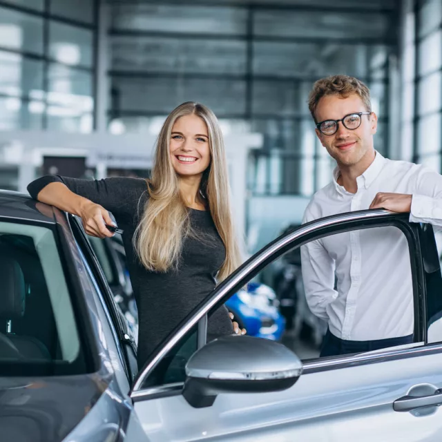 Twee klanten bekijken een nieuwe auto in de showroom van een autodealer, met een vrouw die naast de auto staat en een man die de deur openhoudt.